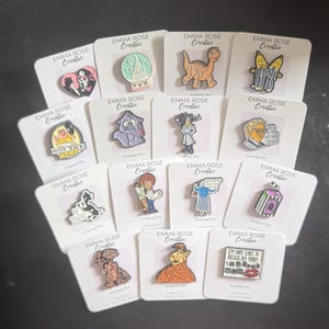 May include: A collection of enamel pins on white cards. The pins feature various designs, including a crystal ball, a dinosaur, and a cartoon character. Each card is labeled "EMMA ROSE Creative".