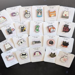 Peut inclure: Une collection d'épingles émaillées avec des motifs de chats variés. Les épingles présentent des chats dans différents styles, certains avec du texte comme "CHONK" et "First of all, I'm a delight." Chaque épingle est présentée sur une carte blanche.