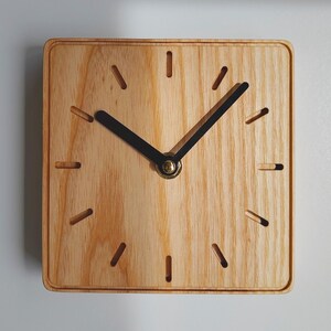 Könnte beinhalten: Eine quadratische Wanduhr aus hellem Holz mit sichtbarer Maserung. Das Zifferblatt verfügt über zwölf eingelassene Schlitze als Stundenmarkierungen und zwei schwarze Zeiger, die an einem zentralen goldfarbenen Metallstift befestigt sind.