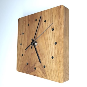 Könnte beinhalten: Quadratische Wanduhr aus Holz mit natürlicher Holzmaserung. Die Uhr hat schwarze Zeiger und schwarze Punktmarkierungen. Die Uhr ist hellbraun und hat ein modernes Design.