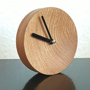 Könnte beinhalten: Eine runde Holzuhr mit hellem Holzton. Sie hat schwarze Zeiger und einen kleinen silberfarbenen Mittelpunkt. Die Holzmaserung ist auf dem Zifferblatt sichtbar. Die Uhr ist ein dekorativer Artikel.