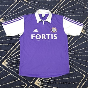 Puede incluir: Una camiseta de fútbol Adidas morada con mangas y cuello blancos. La camiseta presenta la palabra "FORTIS" en letras blancas, el logo de Adidas y un escudo del equipo. Las mangas tienen rayas blancas.