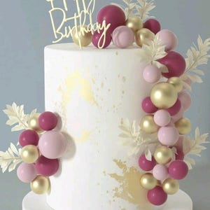 May include: A white birthday cake with gold accents and a "Happy Birthday" cake topper. The cake is decorated with pink, gold, and burgundy spheres, and white leaf accents. The cake sits on a white and gold cake stand.