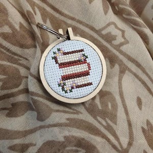 May include: A round, wooden embroidery hoop keychain featuring a cross-stitched design of a stack of books. The books are brown and white, surrounded by small purple and green floral accents on a light blue background.