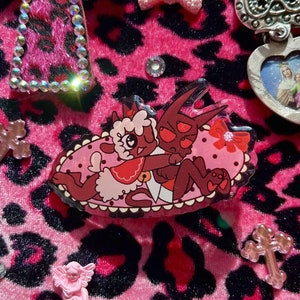 May include: A pink and black leopard print fabric background with a clear acrylic charm featuring a red devil holding a white lamb. The charm is surrounded by pink and white bows, rhinestones, and crosses.