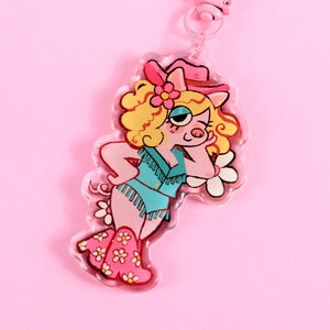 May include: A pink and white pig character wearing a pink cowboy hat and blue denim vest with fringe. The character is wearing pink boots with white flowers. The character is holding a white flower in its right hand. The character is on a clear acrylic keychain.