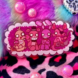 Peut inclure: Un autocollant rectangulaire avec des personnages de dessins animés roses et rouges, encadré d'une bordure festonnée blanche. L'autocollant est posé sur une fausse fourrure rose et violette, une boîte en forme de cœur et des perles irisées.
