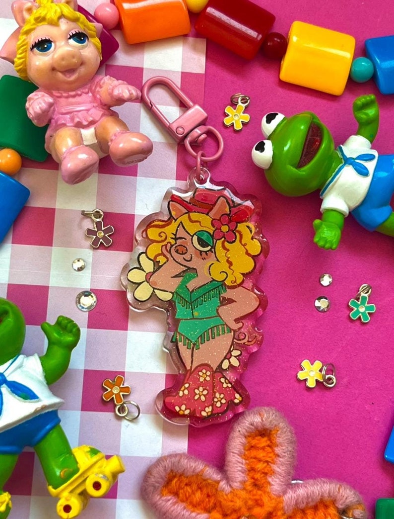 May include: A pink and green pig character keychain with a sparkly green vest and pink boots. The keychain is made of clear plastic and has a pink metal clasp.