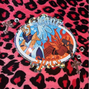 May include: A holographic sticker with a white angel and a red devil, both with crosses on their chests. The sticker says "Sugar Spice" in a dripping font.