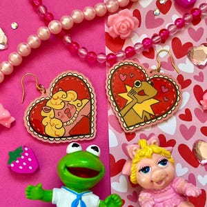 Puede incluir: Pendientes en forma de corazón con personajes de dibujos animados, bordes rojos y fondo rosa. La imagen incluye una figura de rana verde, una figura de cerdo rosa y collares de cuentas rosas. También es visible un dije en forma de fresa.