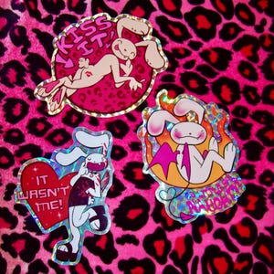 May include: Three holographic stickers featuring a cartoon bunny rabbit in different poses. The first sticker shows the bunny lying on its back with the text "Kiss It!" The second sticker shows the bunny holding a knife with the text "It Wasn't Me!" The third sticker shows the bunny with wings and a devil tail with the text "Fire Me Up, You Naughty Bunny!"