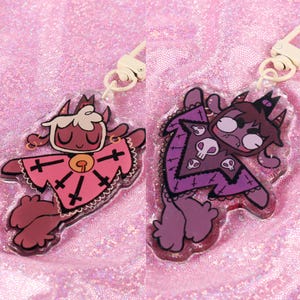 May include: Two colorful acrylic charms featuring cartoon characters with horns and wings. The charms are attached to keychains with gold-colored metal rings. The first charm is pink and red with a cross pattern and the second charm is purple with a skull pattern.