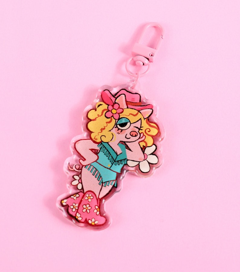May include: A pink and white pig character wearing a pink cowboy hat and blue denim vest with fringe. The character is wearing pink boots with white flowers. The character is holding a white flower in its right hand. The character is on a clear acrylic keychain.