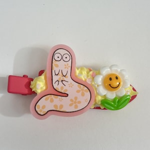 May include: A pink and yellow floral patterned hair clip with a smiling flower and a worm design.