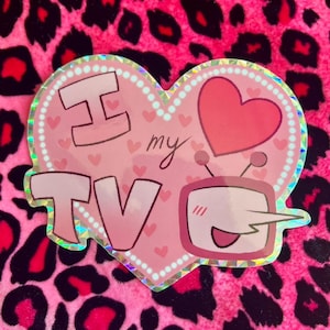 May include: A pink heart-shaped sticker with the words "I love my TV" in a playful font. The sticker features a cartoon TV with a smiling face and a red heart. The background is a pink leopard print.