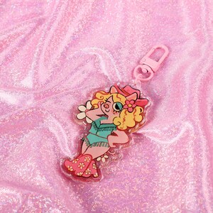 May include: A pink and white pig character wearing a cowboy hat and boots, with a flower in her hair, is featured on a clear acrylic keychain. The keychain has a pink metal clasp.