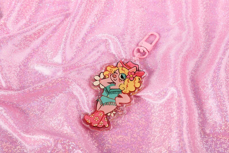 May include: A pink and white pig character wearing a cowboy hat and boots, with a flower in her hair, is featured on a clear acrylic keychain. The keychain has a pink metal clasp.
