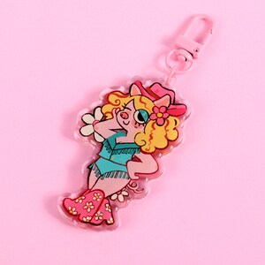 May include: A pink pig wearing a blue and white cowboy outfit with a pink hat and a flower. The pig is holding a flower in its hand. The pig is a keychain charm.
