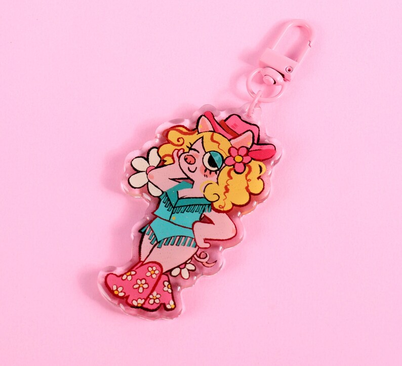 May include: A pink pig wearing a blue and white cowboy outfit with a pink hat and a flower. The pig is holding a flower in its hand. The pig is a keychain charm.