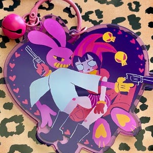 May include: A heart-shaped acrylic keychain featuring a purple background with a pink rabbit character and a human-like figure holding guns. The design includes small yellow skulls and pink hearts. A pink metal ring and a small pink bell are attached.