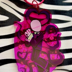 May include: A translucent pink acrylic keychain featuring a character with a pale oval head, wearing a dark suit and tie, surrounded by dark purple tentacles. The design includes a heart-shaped top and a small pink bell attached to the pink ring.