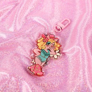 May include: A pink and white keychain featuring a cartoon pig wearing a cowboy hat and a blue and green outfit. The keychain is made of clear acrylic and has a silver metal ring.