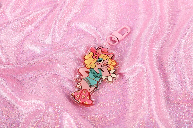 May include: A pink and white keychain featuring a cartoon pig wearing a cowboy hat and a blue and green outfit. The keychain is made of clear acrylic and has a silver metal ring.
