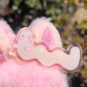 May include: A clear acrylic charm featuring a white, wavy, worm-like character with pink eyes and small arms, attached to a soft, light pink plush fabric base with visible stitched details and a fuzzy texture.