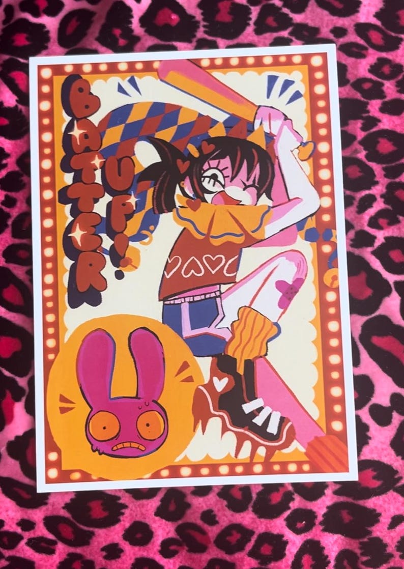 May include: Art print featuring a cartoon illustration of a girl swinging a baseball bat, with the word "Batter" above her. The print has a pink, orange, and blue color scheme, with a cartoon rabbit in the lower left corner.