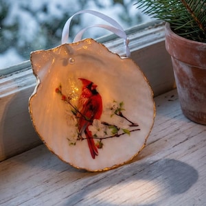 May include: A decorative seashell ornament featuring a red cardinal bird perched on a flowering branch. The shell has a gold-painted edge and a white ribbon for hanging. The background shows a snowy scene and a small potted evergreen.