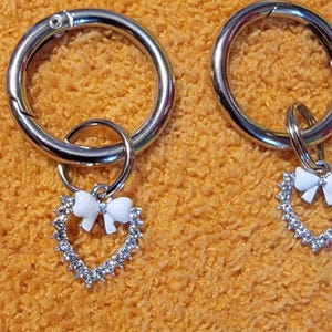 May include: Two silver keychains with a heart-shaped charm with a white bow. The charms are adorned with rhinestones.
