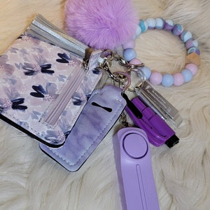May include: A purple and white floral patterned wallet with a silver tassel, a purple keychain with a small purple alarm, a purple and blue beaded bracelet, and a fluffy purple pom-pom.