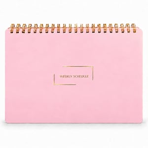 May include: A light pink weekly schedule planner with a gold spiral binding at the top. The front cover features the text WEEKLY SCHEDULE printed in gold, centered within a thin gold rectangular frame with open corners.