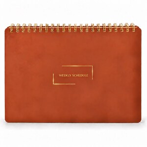 May include: A rust-colored weekly schedule planner with a smooth, matte cover and gold-toned wire-o binding at the top. The center of the front cover features the text WEEKLY SCHEDULE printed in gold, framed by two thin gold rectangular brackets.