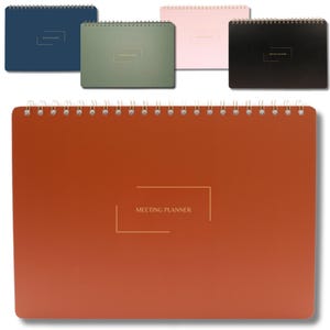 May include: A collection of spiral-bound meeting planners in various colors: navy blue, green, pink, and black. The large, rust-colored planner in the foreground features the text "MEETING PLANNER" in gold lettering. The planners are designed for organizing meetings and schedules.