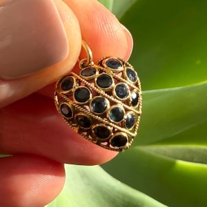 May include: A heart-shaped pendant crafted from yellow gold, featuring a lattice-like wire frame set with numerous small, round, dark blue faceted gemstones. The piece includes a small gold jump ring at the top for attachment to a chain.