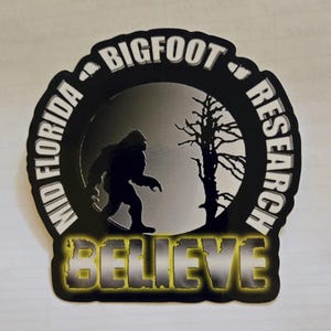 May include: A black and white sticker featuring a silhouette of Bigfoot walking in front of a tree. The sticker has the words "Mid Florida Bigfoot Research" and the word "BELIEVE" in a yellow and black font.