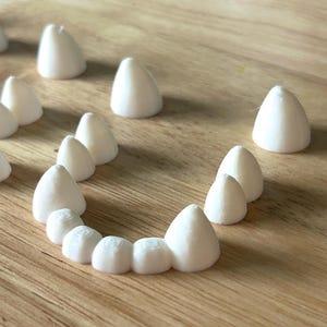 May include: A collection of small, white, cone-shaped objects arranged on a wooden surface. Some are scattered, while others form a semi-circle. The objects have a smooth texture and rounded tips, suggesting a dental or craft theme.