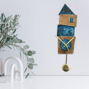Op de afbeelding: Een blauwe en bruine houten klok met een slinger. De klok is gevormd als een huis met een vierkant gezicht en gouden wijzers. De klok hangt aan een witte muur.