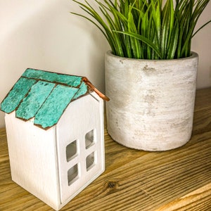 Może przedstawiać: Mały, biały drewniany domek z turkusowym i miedzianym dachem. Dom ma cztery kwadratowe okna i stoi na drewnianej półce obok betonowej donicy wypełnionej zieloną sztuczną trawą. Przedmiot dekoracyjny.