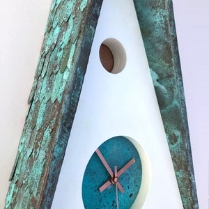 May include: A unique, triangular wall clock with a weathered copper finish. The clock face is teal with copper hands, set within a white frame. The roof has a textured, scale-like pattern. The clock's design is modern and artistic.