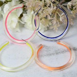 May include: Four pairs of clear hoop earrings with colorful accents. The earrings have a transparent design with neon pink, blue, yellow, and orange inner rims. The earrings are displayed on a light-colored surface with white flowers in the background.