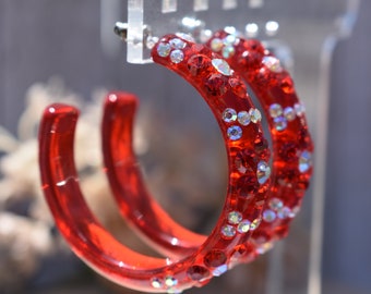 May include: A pair of red hoop earrings with clear rhinestones. The earrings are large and have a shiny finish.