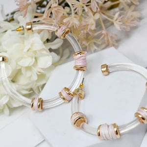 May include: Clear acrylic hoop earrings with gold-tone accents and pink detailing. The earrings have a post back closure and are decorated with pink and gold-tone cylindrical beads. The earrings are displayed on a white surface with floral accents.