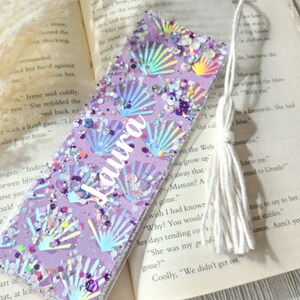 May include: A purple and iridescent glitter bookmark with a seashell pattern and the name "Laura" written on it. The bookmark has a white tassel.