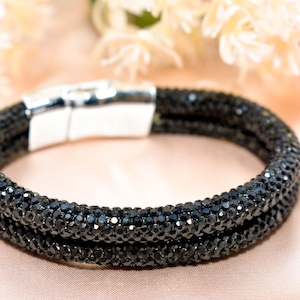 May include: A black beaded bracelet with a magnetic clasp. The bracelet is made of small, dark beads, creating a textured surface. The clasp is silver-colored. The background is a soft, peach-colored fabric with blurred floral accents.