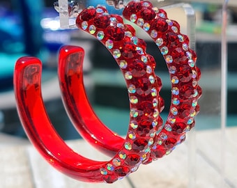 May include: A pair of red hoop earrings with a clear rhinestone design. The earrings are made of plastic and have a shiny finish.