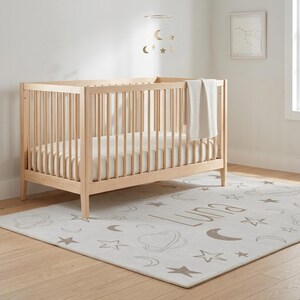 May include: A light wood crib with vertical slats, a white mattress, and a cream-colored blanket. A rug with a celestial design, including stars, moons, and the name "Luna", is on the floor. A mobile with gold moons and stars hangs above the crib.