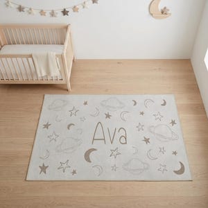 May include: A rectangular, light gray rug with a space theme. The rug features planets, stars, and crescent moons in a muted brown color. The name "Ava" is printed in the center. The rug is placed on a wooden floor.