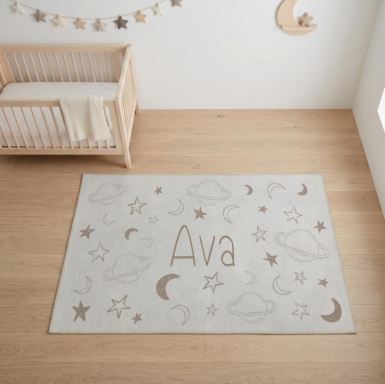 May include: A rectangular, light gray rug with a space theme. The rug features planets, stars, and crescent moons in a muted brown color. The name "Ava" is printed in the center. The rug is placed on a wooden floor.
